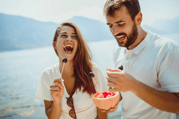 Pareja joven comiendo fruta en la playa- fiesta de verano con un amigo — Foto de Stock