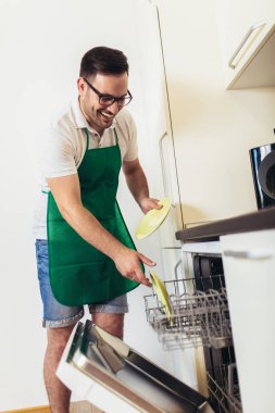 Adam mutfakta ev işi yapıyor, bulaşıkları bulaşıkmakinesine dolduruyor.