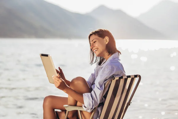 Улыбающаяся женщина сидит на палубе кресло у моря с помощью планшета на — стоковое фото