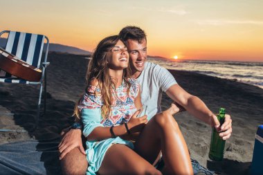 Mutlu genç çift gün batımında bira içip sahilde eğleniyor.
