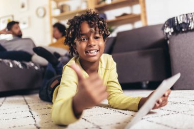 Halıya uzanmış dijital tablet kullanan ve ebeveynleri koltukta oturan bir kız.