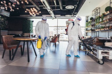 Tehlikeli madde giysileri içindeki profesyonel işçiler kafenin veya restoranın iç kısımlarını dezenfekte ediyorlar, salgın hastalık riski, koronavirüs.