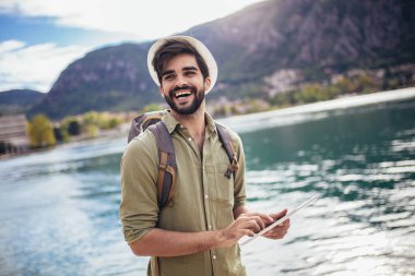 Elinde dijital tabletle arka planda teknelerle turistik bir tatil köyünün limanında yürüyen genç adam..