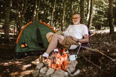 Olgun bir adam çadırın önünde oturuyor ve kamp ateşinin yanında ve bir deftere yazıyor..