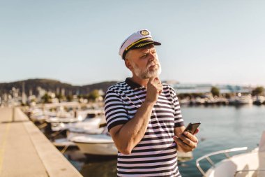 Olgun bir adam denizci gömleği ve şapkası giyip akıllı telefon kullanarak iskelede duruyor..
