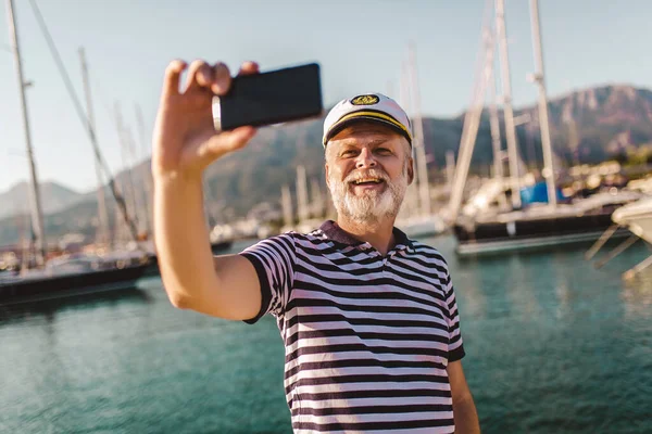 大人の男は 写真を撮るためにスマートフォンを使用して船員のシャツや帽子を身に着けている桟橋に立っています — ストック写真