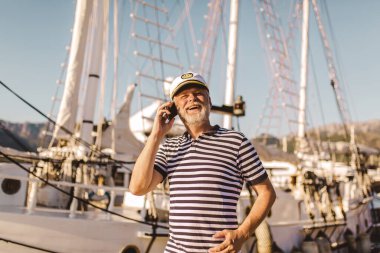 Olgun bir adam denizci gömleği ve şapkası giyip akıllı telefon kullanarak iskelede duruyor..