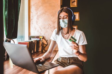 Yüz maskeli sıradan bir kadın, kulaklık, kafede online alışveriş yapmak için dizüstü bilgisayar kartı kullanıyor.