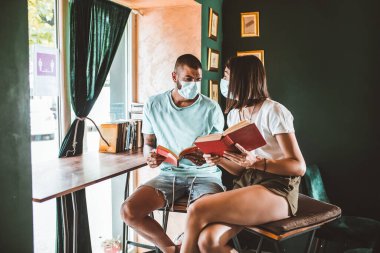 Kahve dükkanında kitap okuyan genç çift koruyucu maskeler takıyor. Virüs koruması.