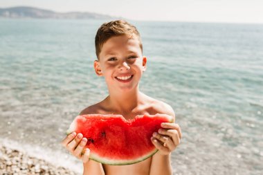 Cheerful child eating juicy watermelon. Kids emotions boy eating watermelon on the background of the sea, the beach, the sea coast. clipart