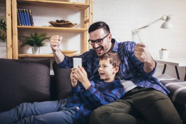 Baba ve oğul evde birlikte vakit geçirirken akıllı telefonlar kullanıyor ve gülümsüyor.