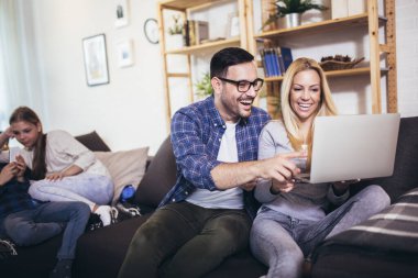 Çocuklar telefon ve dijital tablet kullanarak oynarken, genç ebeveynler evde kanepede dizüstü bilgisayar kullanarak alışveriş yapıyorlar. 