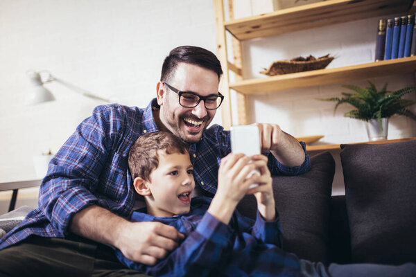 Young father embraces little son family sitting on couch at home using smart phone feels happy cheerful laughing on funny videos.