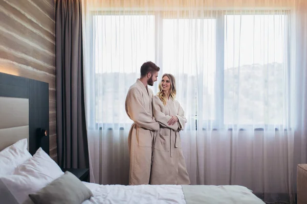 Young happy couple in hotel room in the morning. Just married man and woman standing at window.