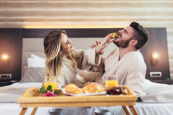 在酒店房间的床上吃早餐的一对微笑的夫妇 — 图库照片