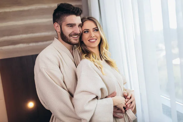 Young happy couple in hotel room in the morning. Just married man and woman standing at window.