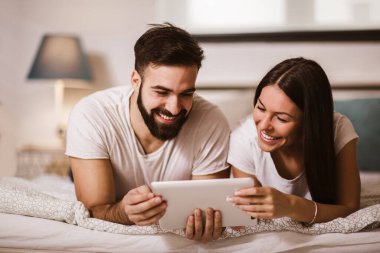 Evde yatakta dijital tablet kullanan gülümseyen rahat genç çift.