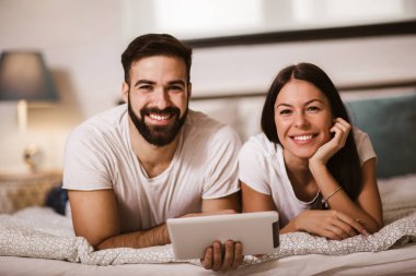 Evde yatakta dijital tablet kullanan gülümseyen rahat genç çift.