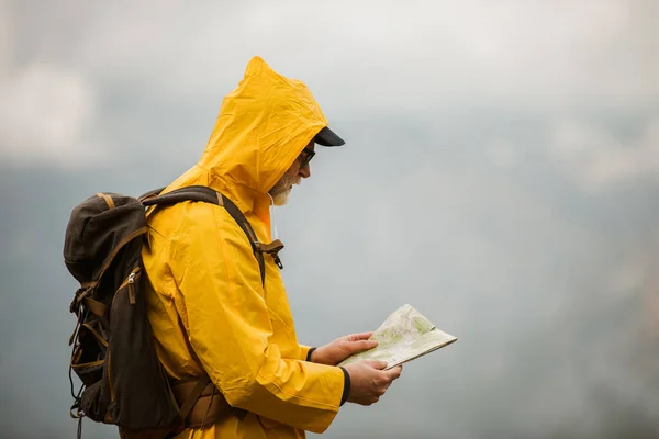 Middle age man traveler in raincoat and backpack using map on hiking