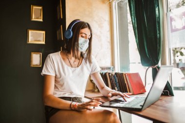Yüz maskesi takan serbest çalışan sıradan bir kadın, kafelerde uzaktan çalışmak için dizüstü bilgisayar kullanan kulaklıklar.