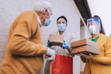 Maskeli genç bayan gönüllüler yaşlı bir adama evinin yakınında yiyecek dolu kutular veriyor. Karantinaya alındı, izole edildi. Coronavirus covid-19. Bağış