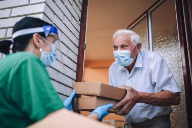 Maskeli genç bir bayan gönüllü yaşlı bir adama evinin yakınında yiyecek dolu kutular veriyor. Karantinaya alındı, izole edildi. Coronavirus covid-19. Bağış