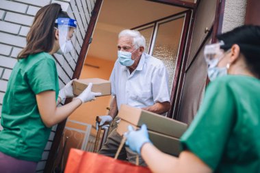 Maskeli genç bayan gönüllüler yaşlı bir adama evinin yakınında yiyecek dolu kutular veriyor. Karantinaya alındı, izole edildi. Coronavirus covid-19. Bağış