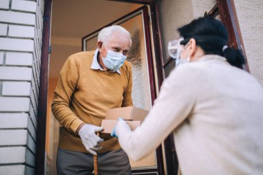 Maskeli genç bir bayan gönüllü yaşlı bir adama evinin yakınında yiyecek dolu kutular veriyor. Karantinaya alındı, izole edildi. Coronavirus covid-19. Bağış