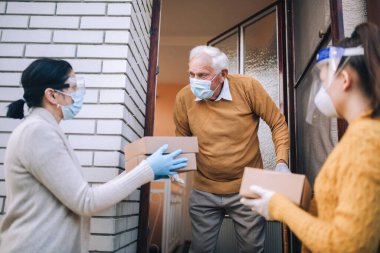 Maskeli genç bayan gönüllüler yaşlı bir adama evinin yakınında yiyecek dolu kutular veriyor. Karantinaya alındı, izole edildi. Coronavirus covid-19. Bağış