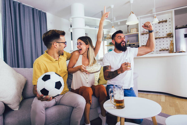 Group of young friends watching a football match at home, cheering and drinking beer.