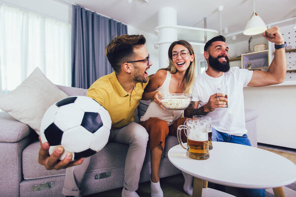 Group of young friends watching a football match at home, cheering and drinking beer.