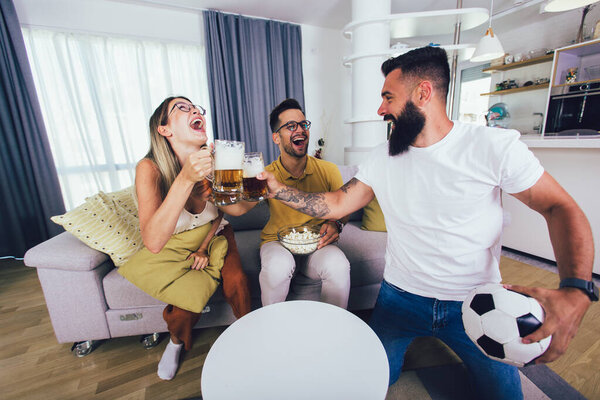 Group of young friends watching a football match at home, cheering and drinking beer.