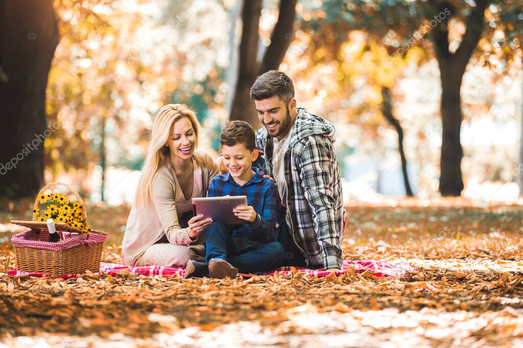 Happy family having picnic in autumn park, using digital tablet.