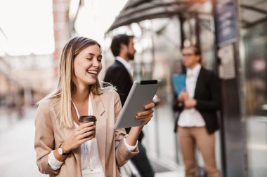 Modern binanın dışına çıkmak için dijital tablet ve kahve taşıyan genç mutlu iş kadını, arka planda iş adamları.