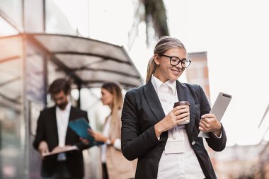 Modern binanın dışına çıkmak için dijital tablet ve kahve taşıyan genç mutlu iş kadını, arka planda iş adamları.