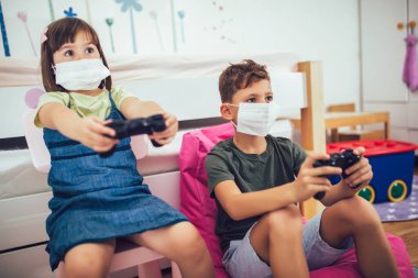 Güzel küçük kız ve oğlan evde koruyucu maske takarak oyun konsolu oynuyorlar.
