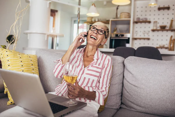 電話で誰かと話している間 ソファに座って ラップトップコンピュータを持つ幸せな中年の女性 — ストック写真