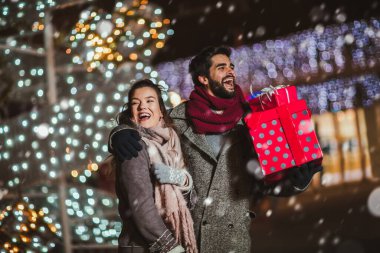 Şehir merkezinde, arka planda tatil ışıkları olan genç bir çift. Noel gecesinde hediye taşıyan bir çift..