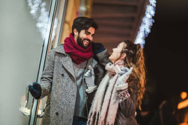 Akşam şehirde yürürken arka planda Noel ışıklarında hediye çantası olan çift. Vitrinlere bak..