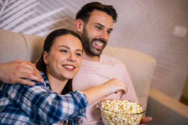 Genç çift oturma odasında uzanıp sarılıyor, televizyon izliyor, patlamış mısır yiyor, meyve suyu içiyor, gülümsüyor ve en sevdikleri dizinin tadını çıkarıyor..