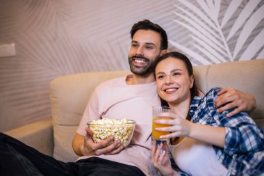 Genç çift oturma odasında uzanıp sarılıyor, televizyon izliyor, patlamış mısır yiyor, meyve suyu içiyor, gülümsüyor ve en sevdikleri dizinin tadını çıkarıyor..