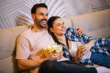 Genç çift oturma odasında uzanıp sarılıyor, televizyon izliyor, patlamış mısır yiyor, meyve suyu içiyor, gülümsüyor ve en sevdikleri dizinin tadını çıkarıyor..