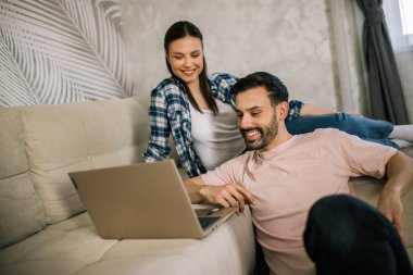 Genç bir çift oturma odasındaki koltukta dinleniyor, birlikte dizüstü bilgisayar kullanıyor, geziyor, alışveriş yapıyor ya da evden çalışıyor..