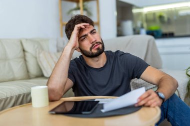 Oturma odasında sakallı genç bir adam faturalarla oturuyor. Günlük mali baskılarla boğuşurken endişeli görünüyor..
