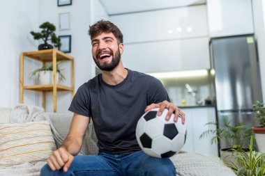 Genç bir adam oturma odasında oturuyor, futbol maçı sırasında tezahürat yapıyor. Bir futbol topu tutarak, en sevdiği takım için heyecan ve tutku gösteriyor..