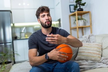 Oturma odasında oturan genç bir adam, bir basketbol maçına kendini kaptırmış, elinde basketbol topuyla maçın heyecanından zevk alıyor..