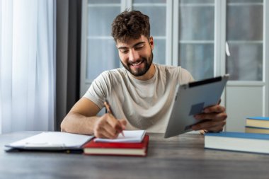 Kitaplarla çevrili bir masada oturan genç bir öğrenci, tamamen tabletindeki sınava çalışmaya odaklanmış, notlar ve ders kitaplarıyla çalışırken kendini adamış..