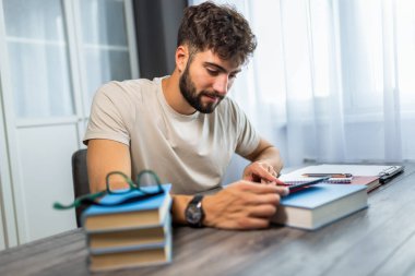 Kitaplarla çevrili bir masada oturan genç bir öğrenci, tamamen tabletindeki sınava çalışmaya odaklanmış, notlar ve ders kitaplarıyla çalışırken kendini adamış..