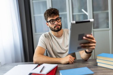 Gözlüklü genç bir öğrenci kitaplarla dolu bir masada oturuyor, tabletindeki sınava çalışmaya odaklanmış, notlarla çalışırken kendini adamış..