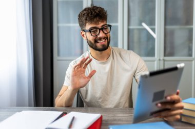 Gözlüklü genç bir öğrenci kitaplarla bir masada oturuyor, online bir derse katılıyor ve bir profesörle tabletinde video aracılığıyla konuşuyor, tamamen öğrenmeye odaklanıyor..
